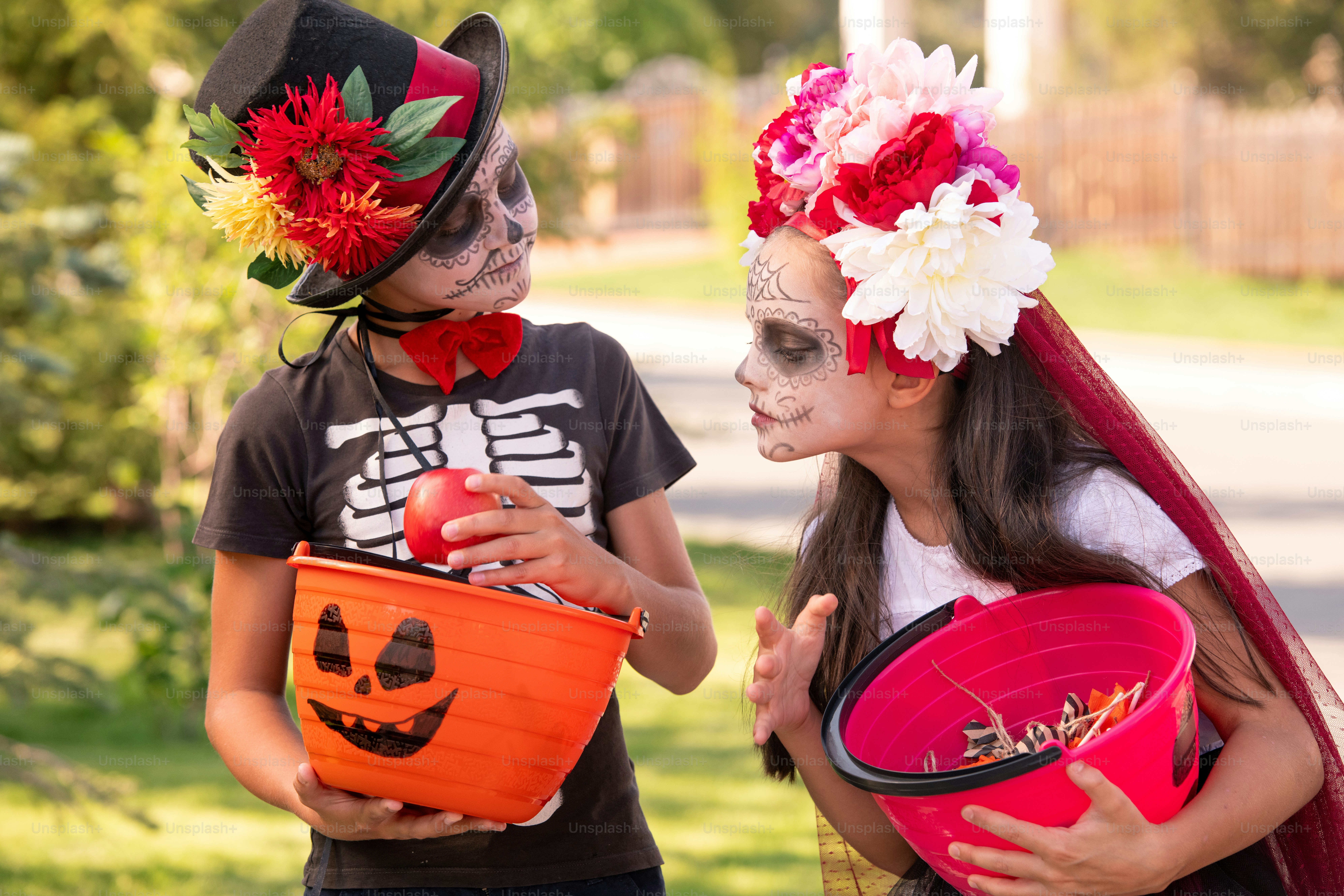 Ragazza halloweeen carina con capelli lunghi scuri che guarda il cestino con dolcetti nelle mani del suo amico che tiene la mela matura rossa mentre si vanta con esso