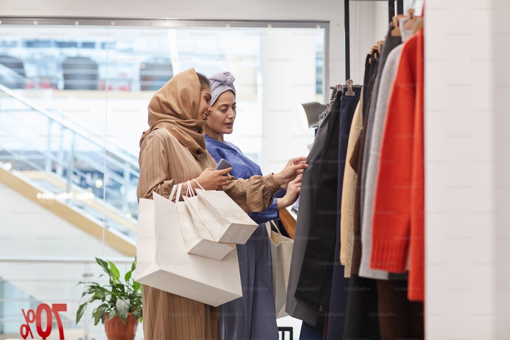 Retrato de vista lateral de dos mujeres jóvenes de Oriente Medio eligiendo ropa mientras disfrutan de las compras en el centro comercial, copie el espacio
