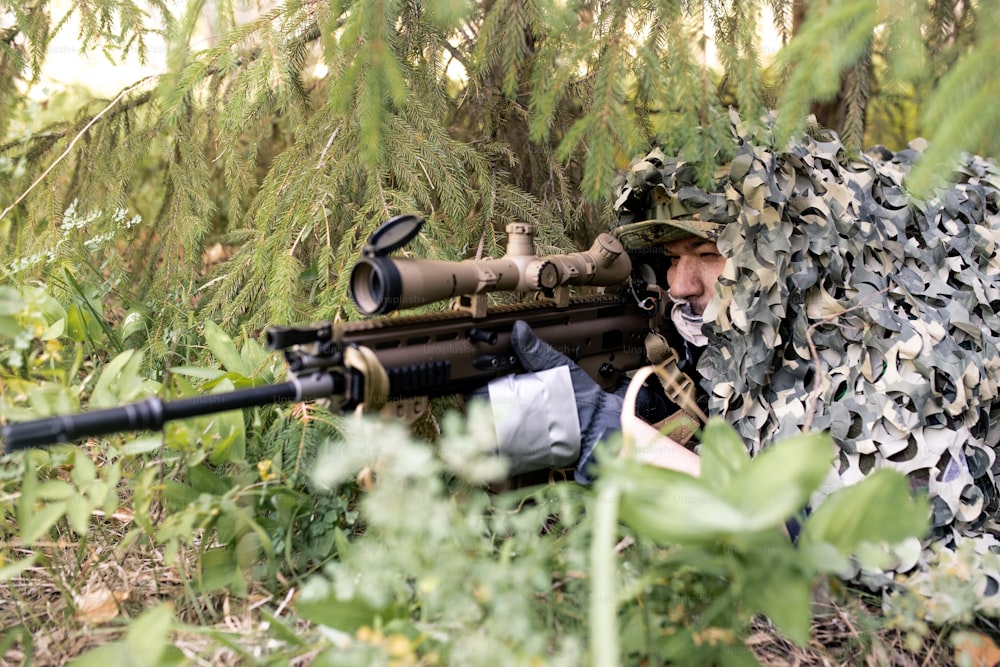 Francotirador concentrado tendido bajo una red de camuflaje en el suelo y mirando a través del visor del rifle