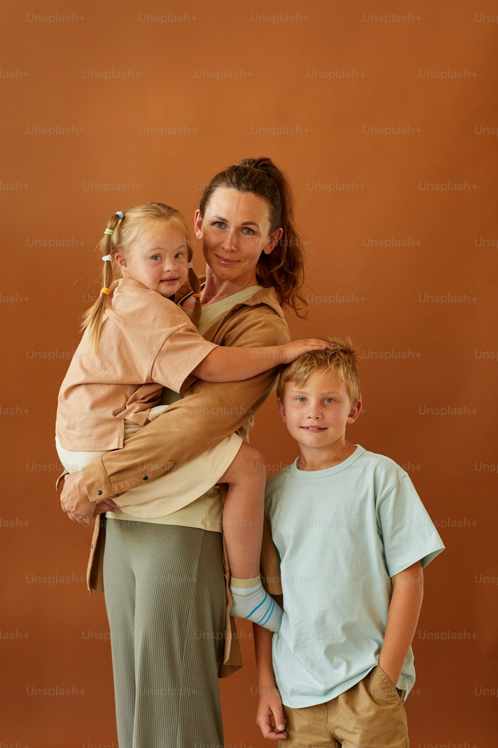 Retrato vertical de una madre madura feliz sosteniendo a su hija con síndrome de Down y un hijo sonriente mientras está de pie contra un fondo marrón liso en el estudio