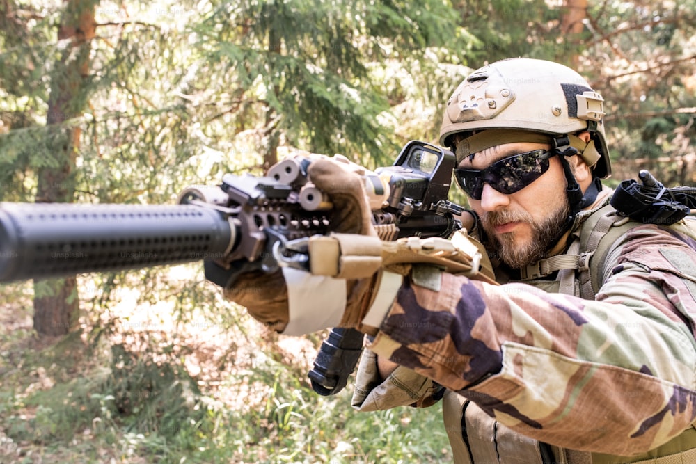 Atirador barbudo de óculos escuros e capacete focado no alvo olhando através de mira de fuzil em floresta