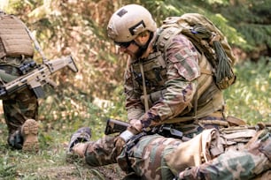 Mann mit Helm und Sonnenbrille untersucht das Bein eines verwundeten Soldaten auf dem Schlachtfeld