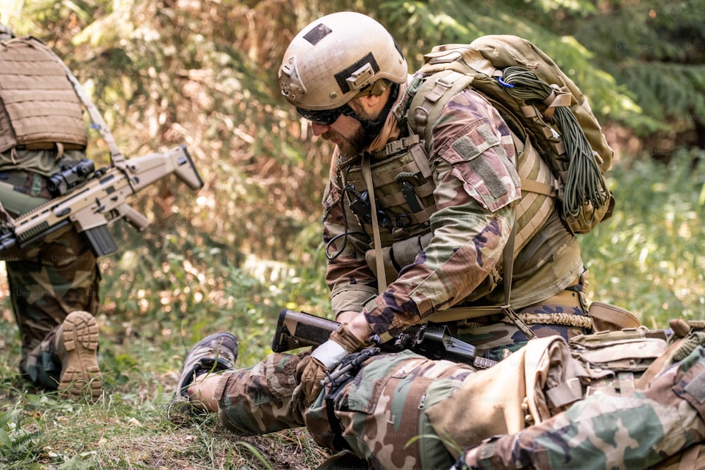 Homem de capacete e óculos escuros examinando perna de soldado ferido no campo de batalha