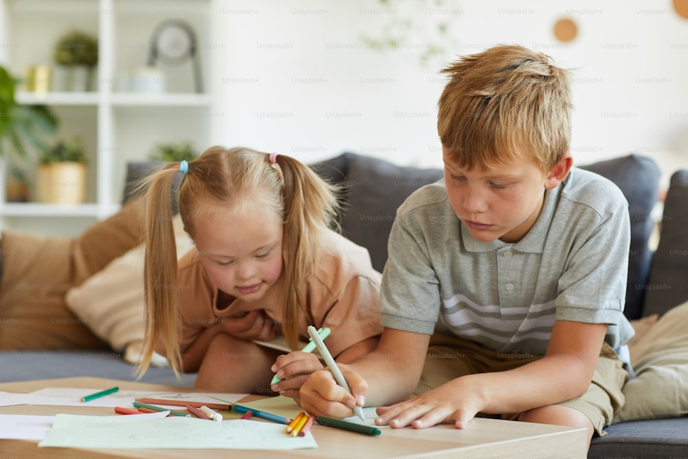 Portrait of cute blonde girl with down syndrome drawing together with older brother at home, copy space