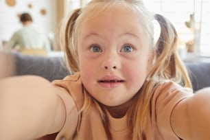 Close up portrait of cute little girl with down syndrome taking selfie and holding camera at home, copy space