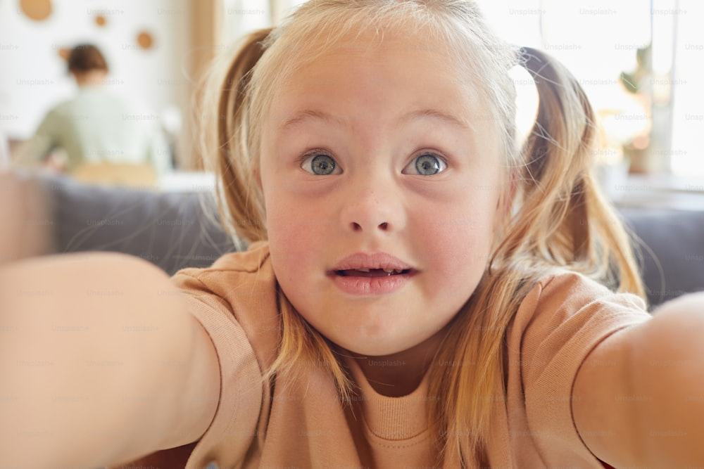 Ritratto ravvicinato della bambina carina con sindrome di Down che scatta selfie e tiene la macchina fotografica a casa, copia lo spazio