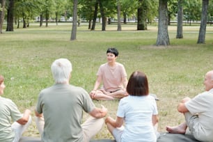 Professional yoga coach sitting in lotus position in park and explaining basics of yoga to senior students