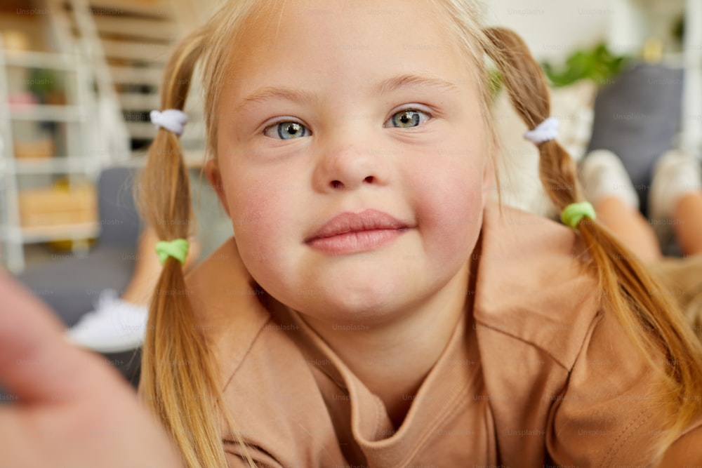 Close up portrait of blonde girl with down syndrome looking at camera while taking selfie photo at home