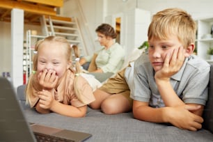 Portrait d’une jolie fille atteinte du syndrome de Down souriant joyeusement en regardant l’écran d’un ordinateur portable et allongée sur le canapé avec son frère à la maison
