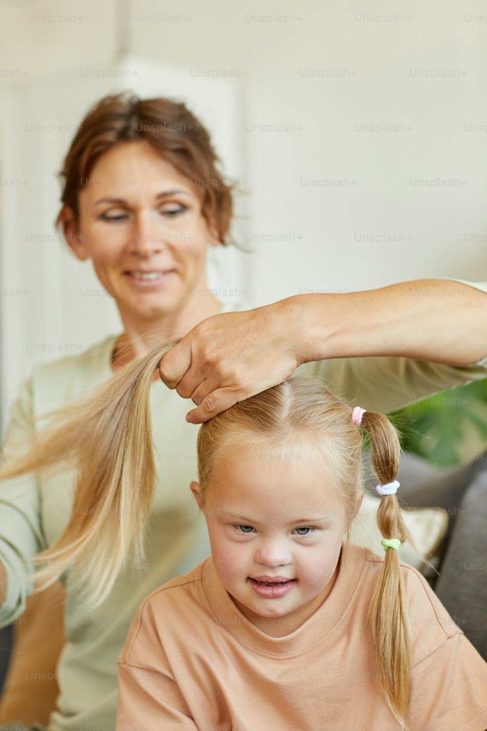 Vertical portrait of smiling mother combing hair of cute girl with down syndrome and tying it in pigtails