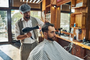 Enthusiastic hair-stylist wearing a fancy hat and blow-drying his client hair after washing it