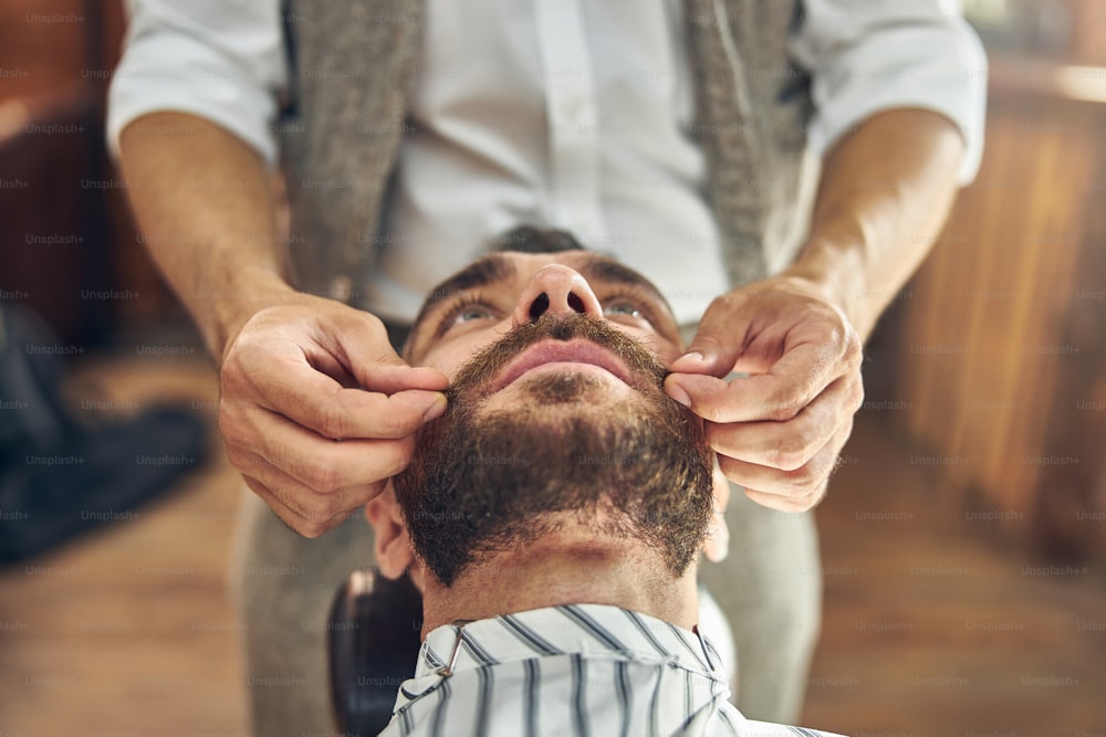 Foto cortada de um cliente barbudo de uma barbearia tendo seu bigode encerado e estilizado por um cabeleireiro