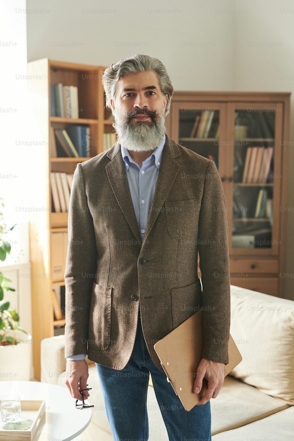 Medium long shot portrait of stylish mature man working as psychologist standing in his office holding clipboard and eyeglasses looking at camera