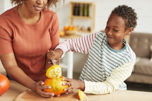 Cheerful African American boy cutting off pumpkin crown. his mother helping him to keep vegetable still