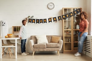 Modern young mother and her son hanging garland with Trick Or Treat lettering on wall for Halloween party