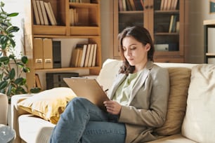 Mulher jovem atraente sentada no sofá na sala de escritório moderna segurando a prancheta fazendo anotações