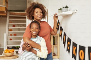 Retrato medio horizontal de una mujer adulta joven feliz y su alegre hijo sonriendo a la cámara mientras decoran la habitación para Halloween
