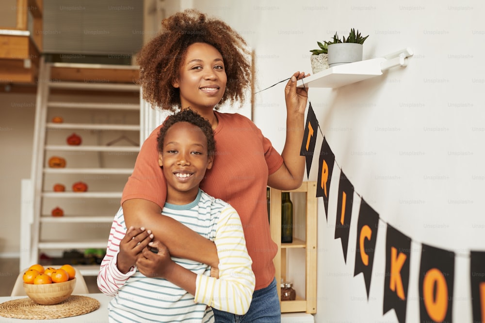 Horizontal medium portrait of happy young adult woman and her cheerful son smiling at camera while decorating room for Halloween