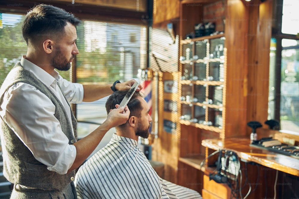 Dedicated young barber looking at his client in the mirror while grooming and styling his hair