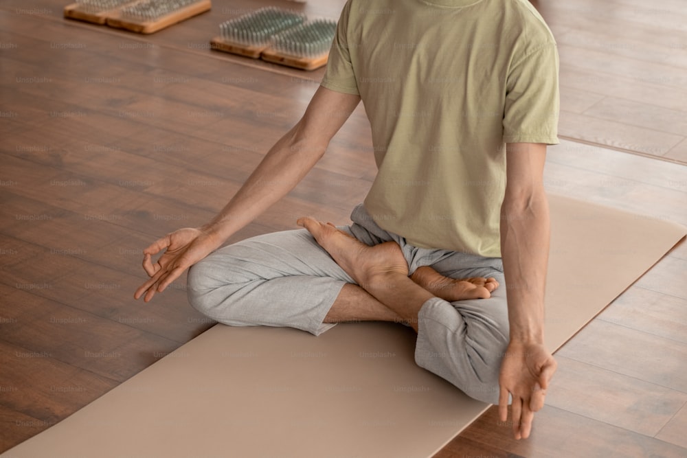 Young man in sportswear keeping balance while sitting on mat in pose of lotus and practicing meditation exercises in home environment