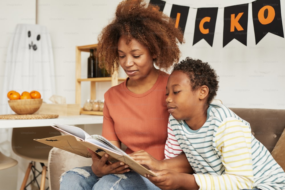 Mulher afro-americana bonita sentada no sofá na sala de estar com seu filho pré-adolescente lendo interessante livro de histórias de Halloween