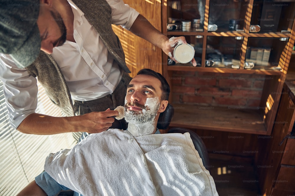 Concentrated well-dressed barber generously applying shaving cream on face and neck of his customer