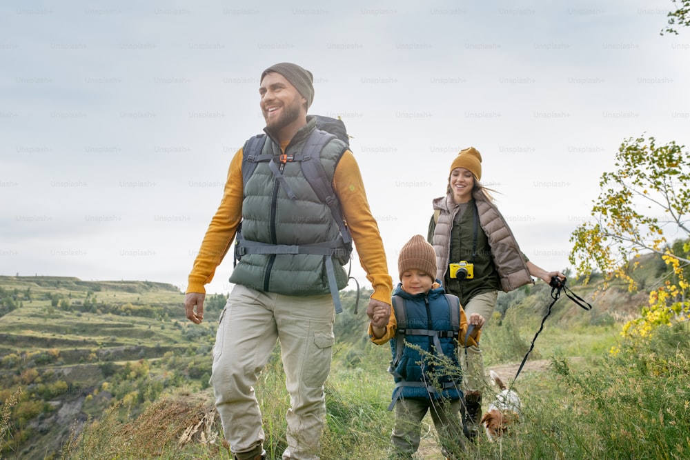 Three happy backpackers in warm casualwear and their cute pet moving down footpath while enjoying weekend trip in natural environment