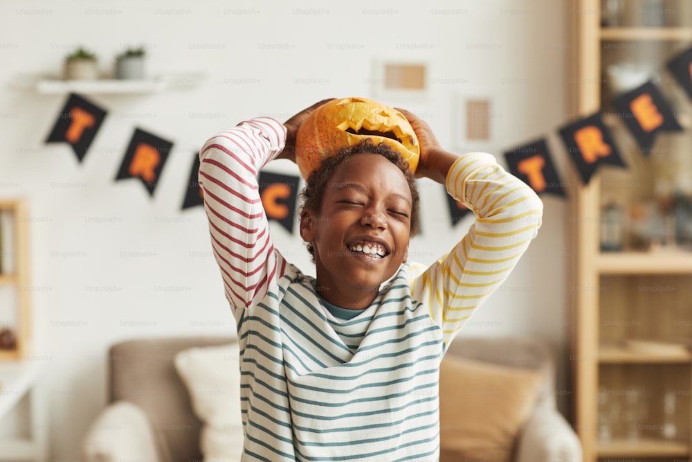 Playful African American boy having fun with Jack O' Lantern pumpkin putting it on his head, medium portrait shot