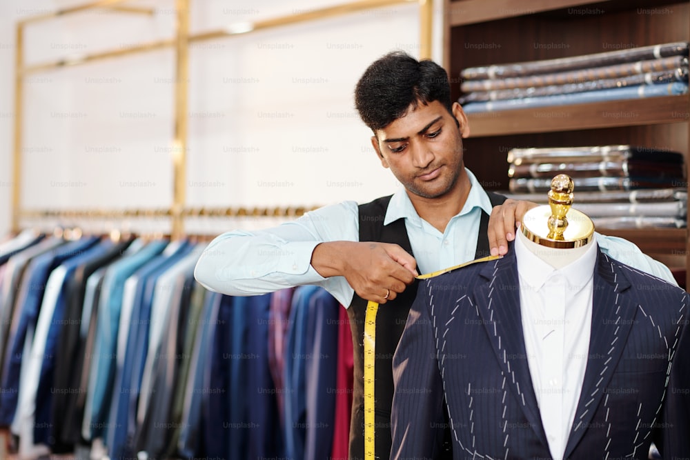 Serious Indian tailor taking measurements of bespoke suit jacket on mannequin