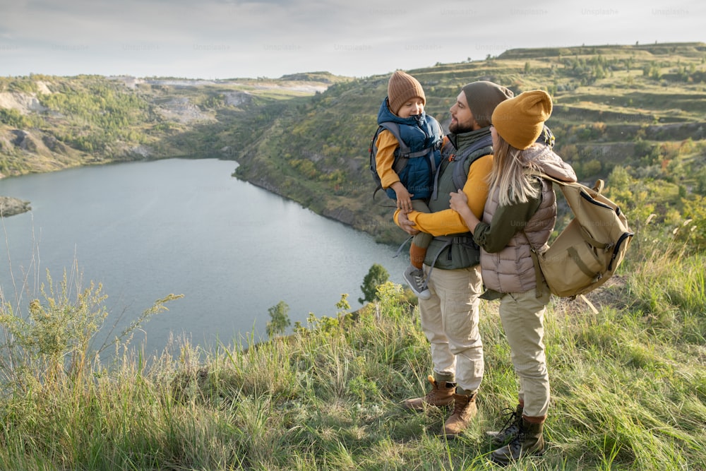Young parents and their little son with backpacks standing on green grass against lake or river surrounded by mountains while enjoying trip
