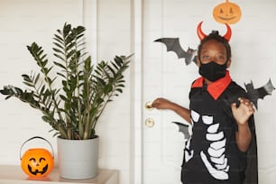 Garçon afro-américain moderne portant un costume de diable avec des cornes rouges et un masque noir sur le visage disant au revoir à ses parents et allant tromper ou traiter avec des amis.