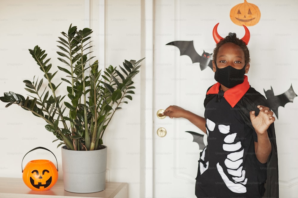 Modern African American boy wearing devil costume with red horns and black mask on face saying bye to his parents and going to trick or treat with friends..