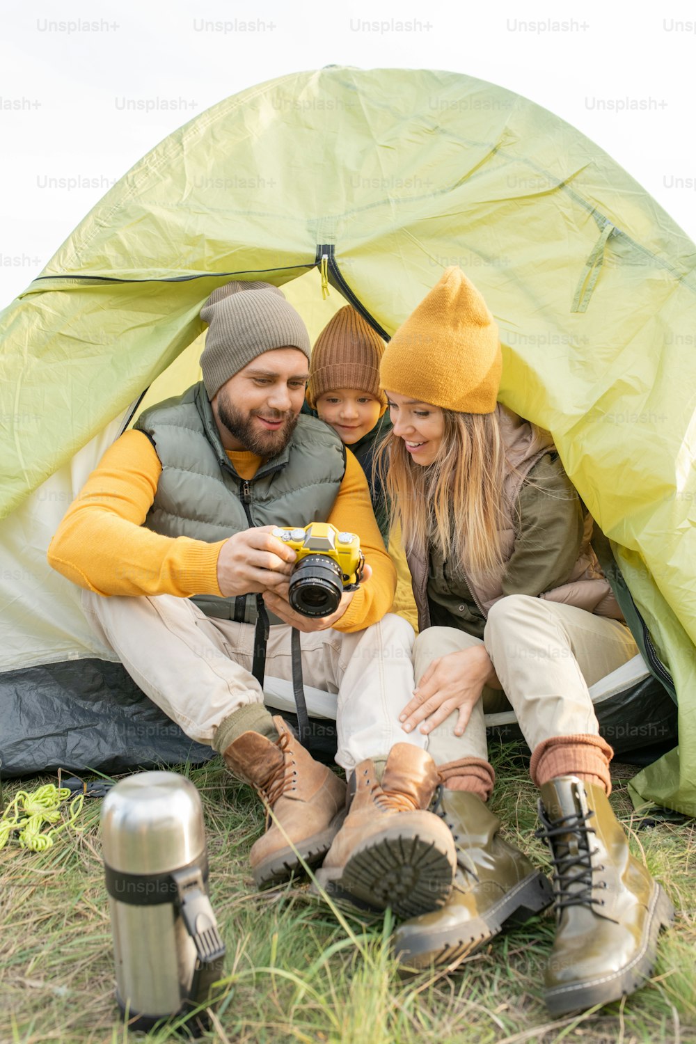 Young man showing photos in photocamera to his son and wife while having rest by tent in natural environment on autumn weekend