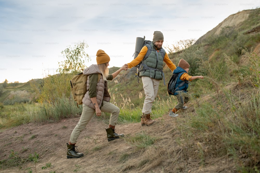 Young active family of hikers with backpacks moving upwards while bearded man helping his wife and son during trip on weekend