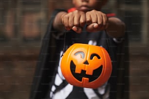 Garçon méconnaissable portant un costume d’Halloween moderne démontrant un panier en plastique Jack O 'Lantern pour les bonbons tout en faisant un tour ou un traitement