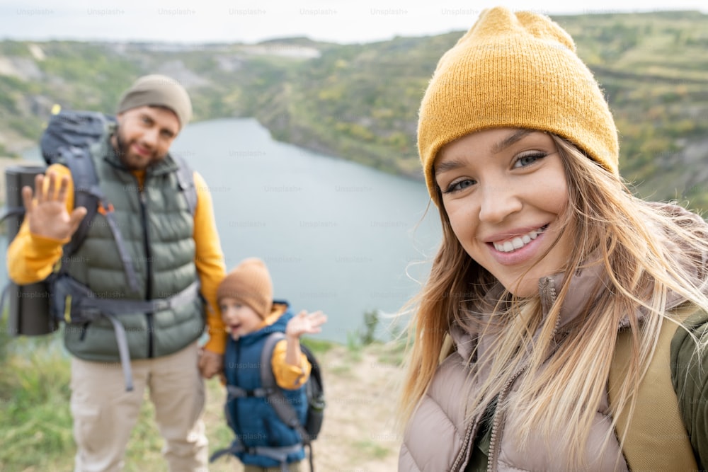 Happy young blond woman with toothy smile making selfie with her husband and cute little son on background during trip in autumn