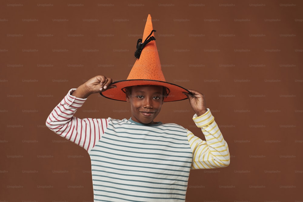 Studio portrait of cheerful young boy standing against brown wall background trying on funny orange wizard hat looking at camera