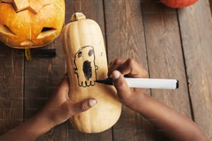 Criança afro-americana irreconhecível desenhando fantasma na cabaça usando caneta de marcador preto para decoração de casa de Halloween, tiro de cima para baixo