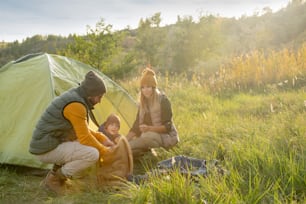 Giovane che prende il cibo dallo zaino mentre è accovacciato in tenda con sua moglie e suo figlio che vanno a riposarsi e pranzare durante il viaggio del fine settimana