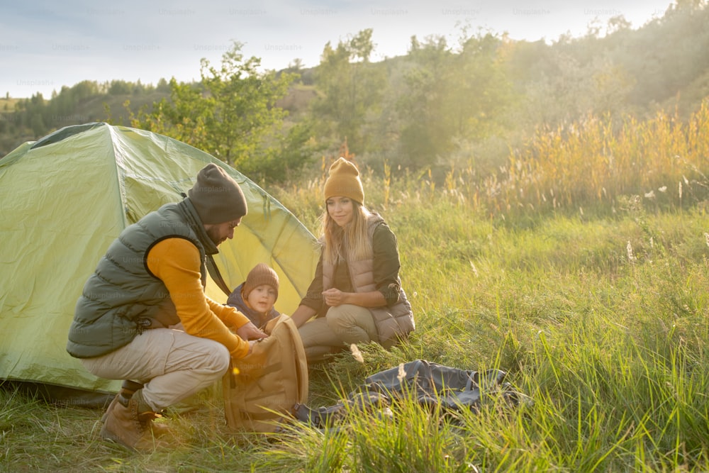 Junger Mann, der Essen aus dem Rucksack nimmt, während er mit seiner Frau und seinem Sohn im Zelt hockt, um sich während des Wochenendausflugs auszuruhen und zu Mittag zu essen