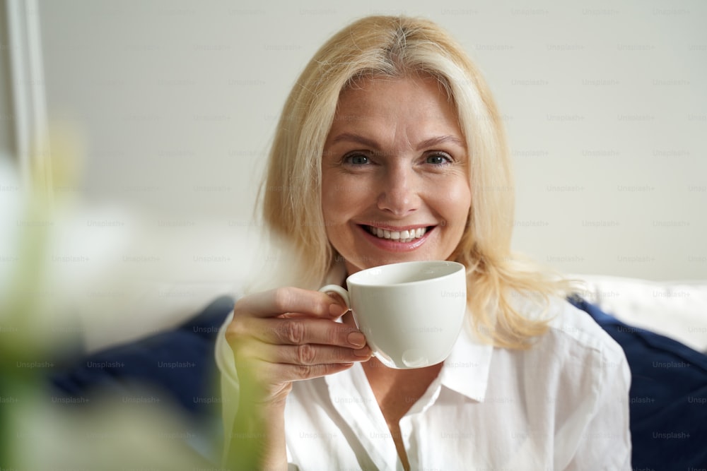 Smiling beautiful adult female holding cup of coffee while looking at camera