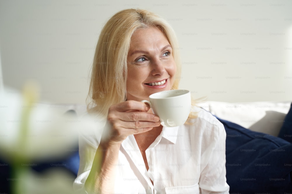 Happy beautiful adult woman holding cup of coffee while looking away indoors