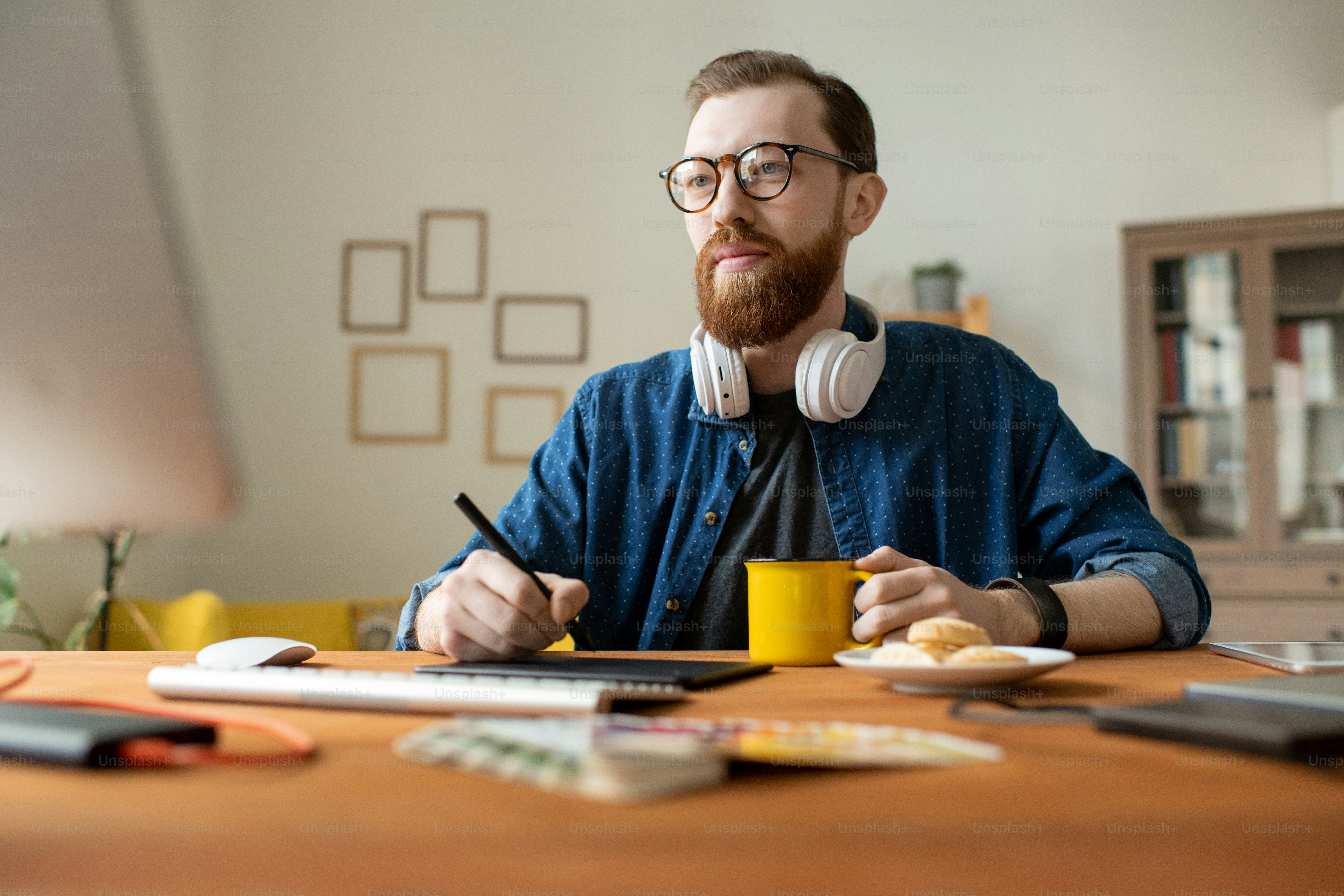 Young creative male freelance designer having drink while holding stylus over tablet and drawing sketches of new logo or website page