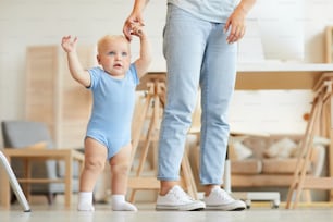 Horizontal shot of unrecognizable mother holding little hand of her cute kid trying to walk at home