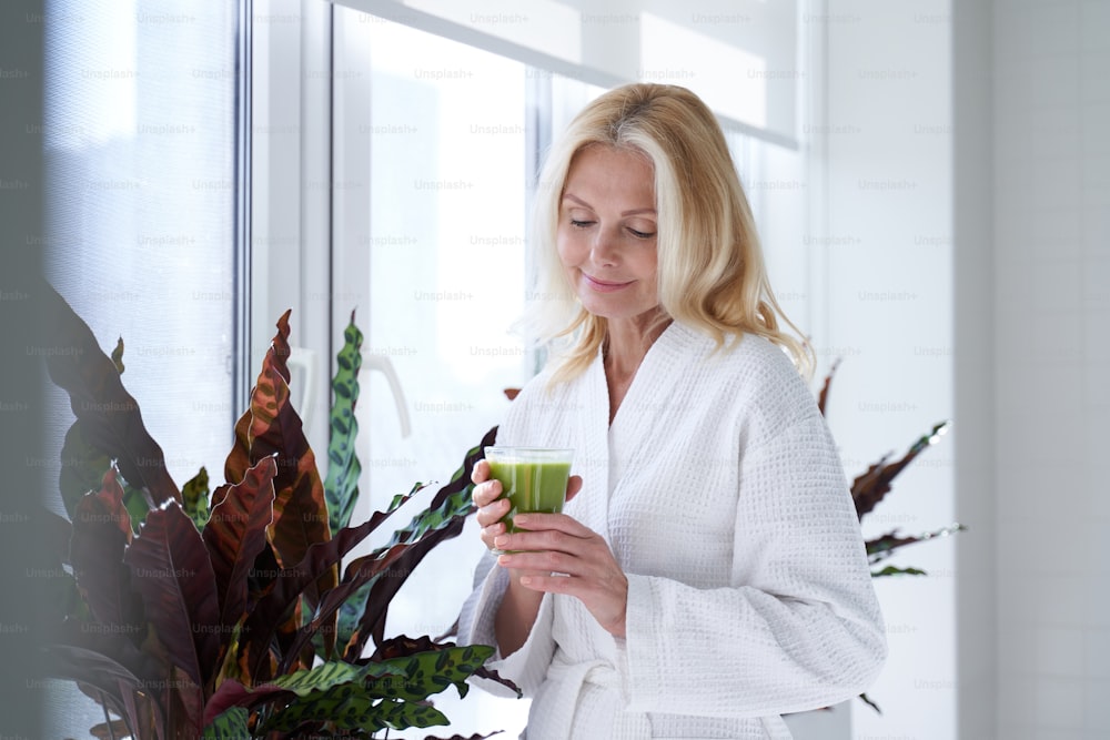 Waist up of smiling beautiful adult lady in white bathrobe and going to drink green smoothies while staying in spa clinic