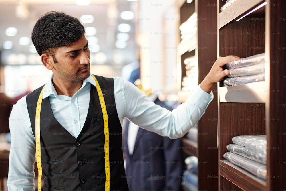 Serious young Indian tailor looking at packed finished clothing items, orders on shelves