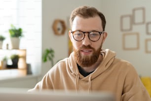 안경을 쓴 젊은 수염 난 프리랜서 디자이너가 원격 근무 중에 온라인 정보를 스크롤하면서 노트북 디스플레이를 보고 있다