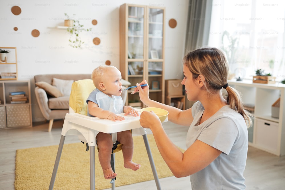 Modern young adult woman feeding her cheerful baby son with porridge in living room at home