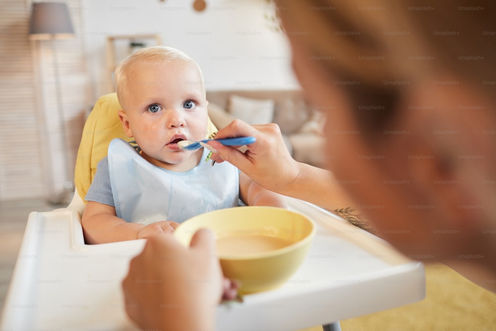 Unrecognizable loving mother feeding her little baby son with tasty fruit squash, over the head shot