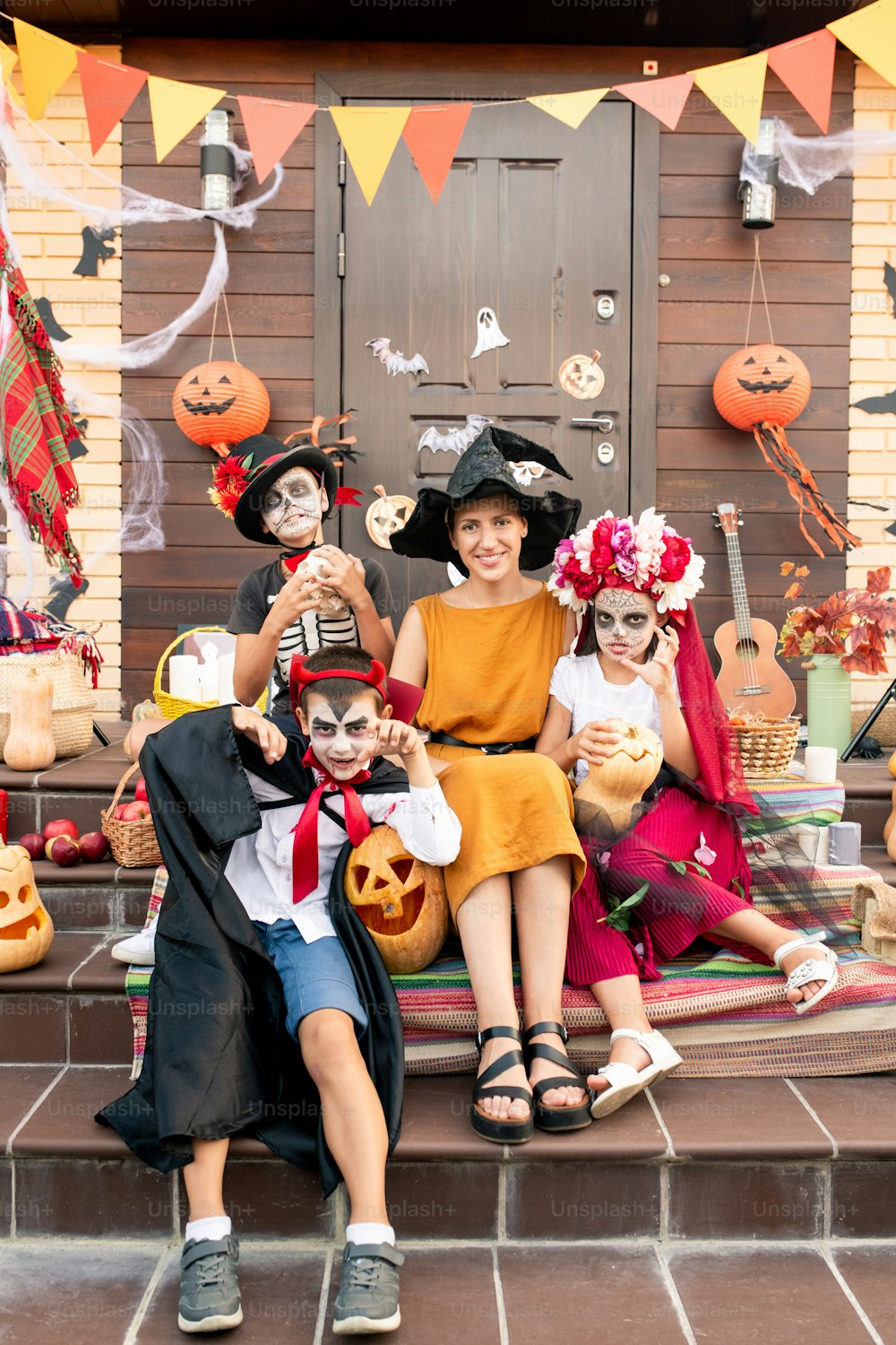 Young happy woman in witch hat and spooky halloween kids in smart costumes sitting on staircase by porch and decorated door of house
